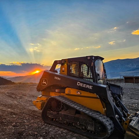 A John Deere 333G Compact Track Loader sitting in the foreground with the sunrise in the background.