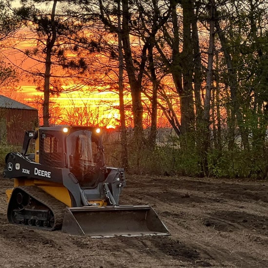 John Deere 323E CTL parked in front of a setting sun.