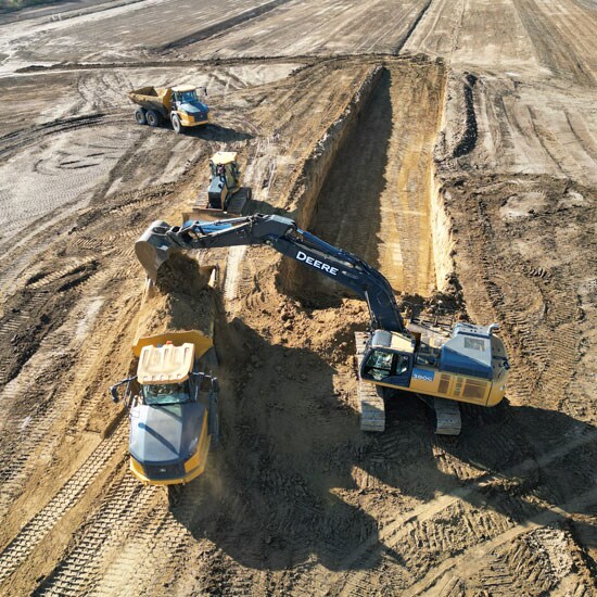 John Deere 380G LC Excavator filling an Articulated Dump Truck while dozer sits behind the articulated dump truck and another articulated dump truck turns to join the line.