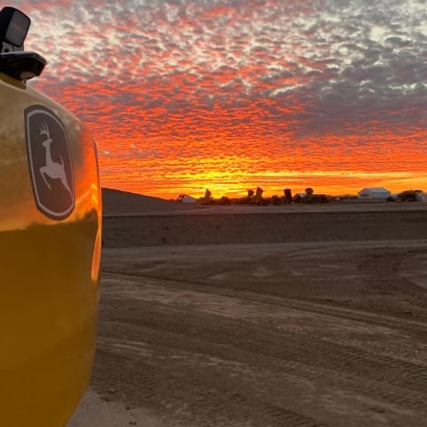 A John Deere excavator is sitting in a dirt field in front of a farm with a sunset in the background.