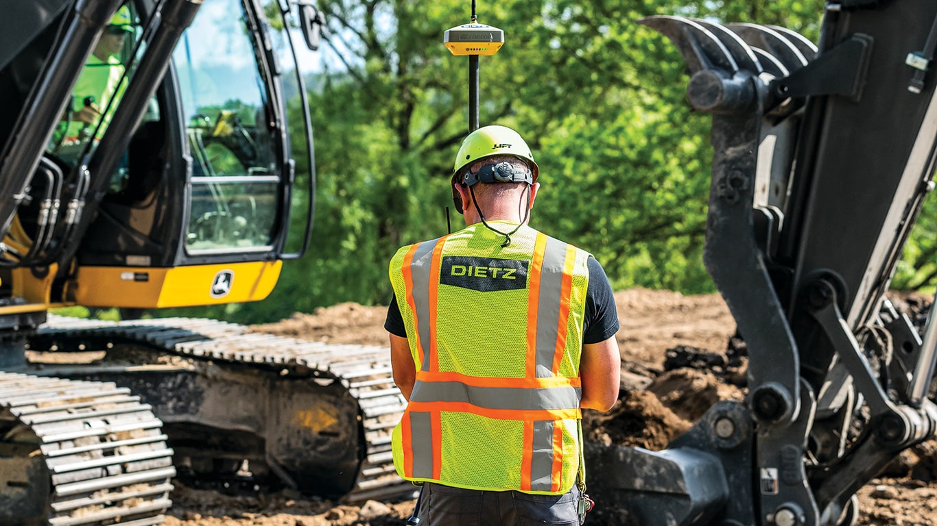 Joe Dietz using a GPS rover with a satellite base station near the John Deere 210G LC Smartgrade Excavator.