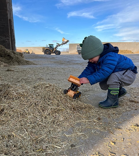 Child playing with a toy replica of a John Deere wheel loader, with the real-life version at work in the background.