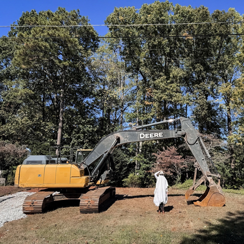 A John Deere 210G LC Excavator sitting in front of trees with a person dressed as a ghost under the boom arm.