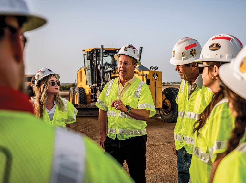 Crew members of Ajax Paving stand near a John Deere 672GP Motor Grader as they discuss the plan of action for the coming day. 