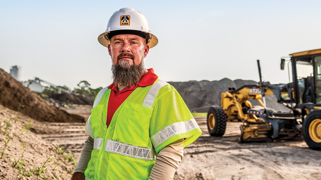 Matt Ohley, operations manager at Ajax Paving, stands on a jobsite with a 672GP Motor Grader in the background.