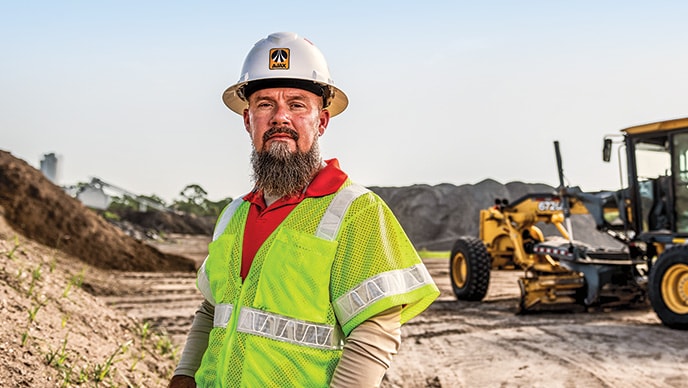 Matt Ohley, operations manager at Ajax Paving, stands on a jobsite with a 672GP Motor Grader in the background. 