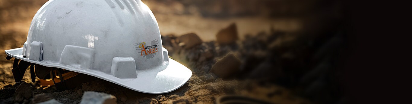 A white construction hard hat with a sticker of the Construction Angels logo on its front lays on the dirt.