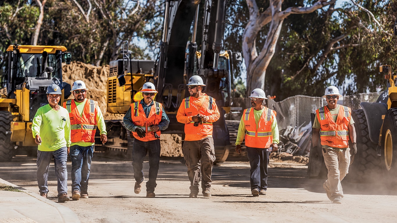 Leonard, Rogeriano, Saul, Ruben, Hermilo, and Jose Luis walk together in front of a parked John Deere excavator and wheel loader and share a conversation at the end of the workday.