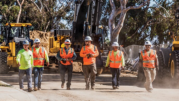 Burtech Pipeline employees Leonard, Rogeriano, Saul, Ruben, Hermilo, and Jose Luis walk together in front of a parked John Deere excavator and wheel loader and share a conversation at the end of the work day. 