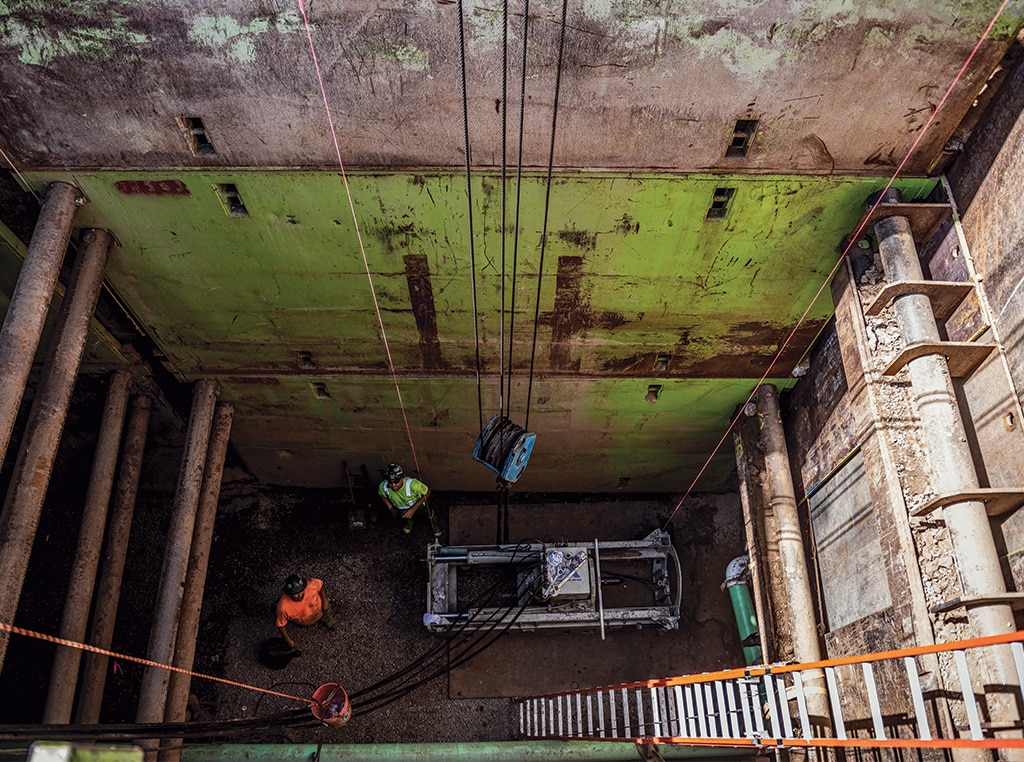 A pair of employees peer up from the shaft as materials are lowered for them on a jobsite.