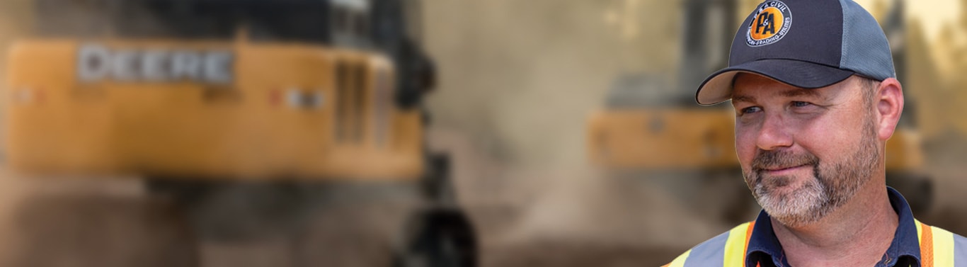 Person in hard hat and safety vest stands at construction site with heavy machinery in the background.