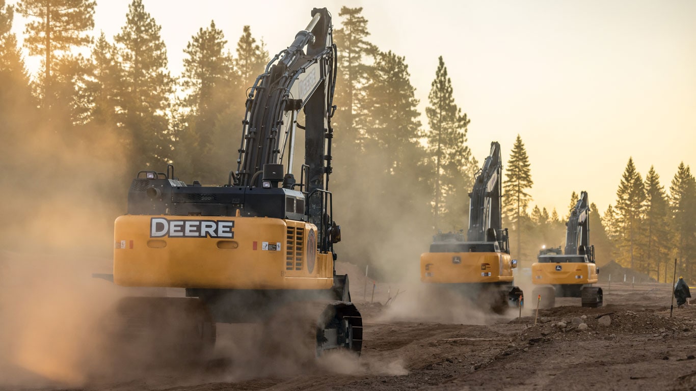 Three excavators work along a dirt roadway, raising dust amid pine trees in warm evening light.