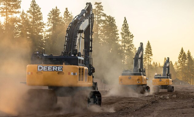 Three excavators work along a dirt roadway, raising dust amid pine trees in warm evening light.