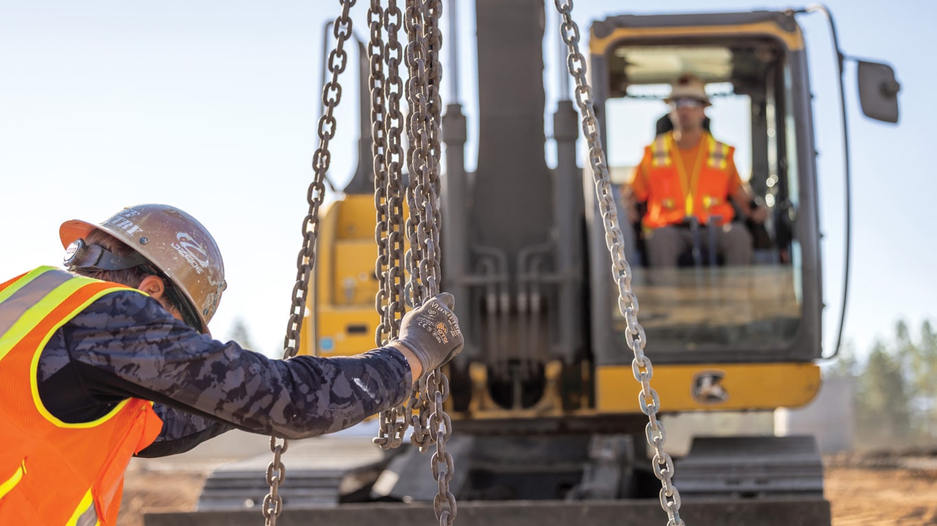 Construction worker in safety vest handles heavy chains beside an excavator at an active job site.