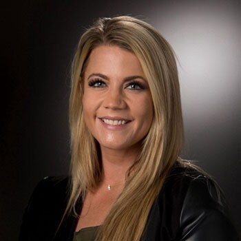 A woman with long blonde hair, wearing a black jacket, poses against a soft-focus dark background.