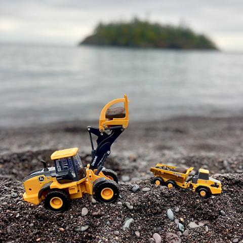 A John Deere 544 P-Tier Wheel Loader and John Deere 460E-II Articulated Dump Truck toy positioned on a dark pebble beach, creating a "sandbox quarry".