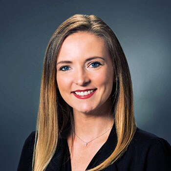 A woman with long, straight brown hair is wearing a black blouse and a delicate necklace, set against a muted gray background.