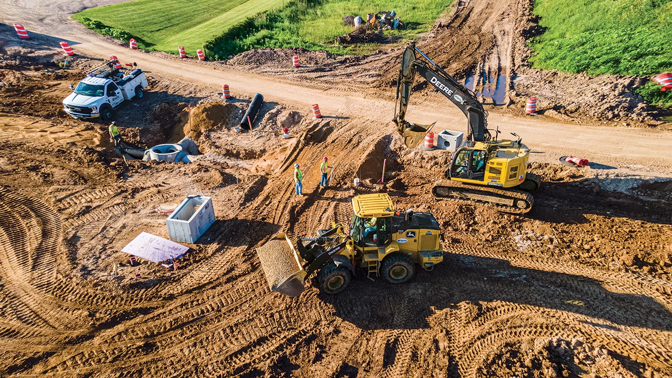 A John Deere 624 P-Tier hauls gravel to an exposed manhole and a 245 G-Tier Excavator fills a trench.