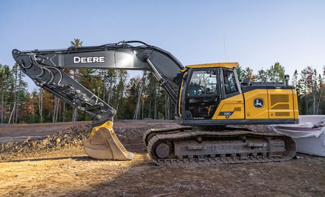 John Deere tracked excavator parked at a construction site with barriers and trees in the background.
