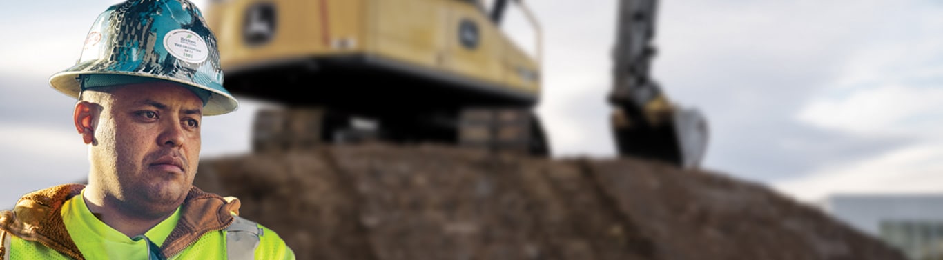 Construction worker in hard hat and safety vest stands at job site with excavator in background.