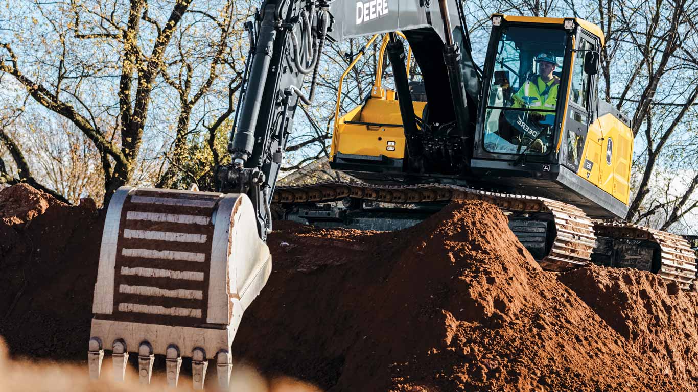Excavator moving a large mound of soil at an outdoor construction site surrounded by trees