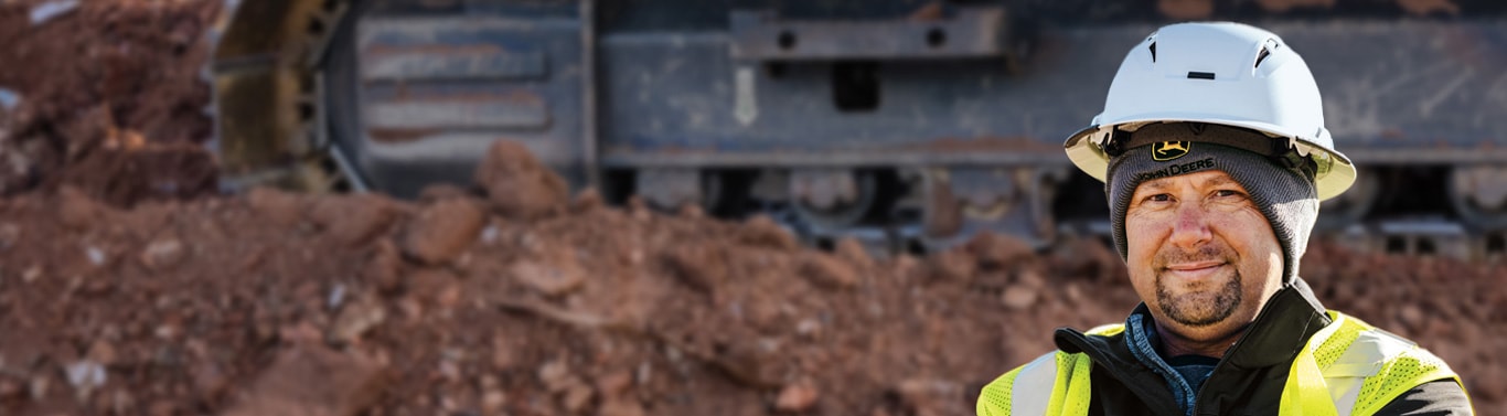 Person wearing a hard hat and high-visibility vest standing at a construction site with equipment behind.