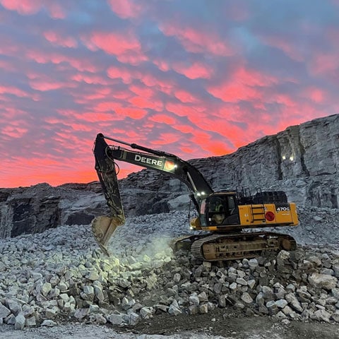 A John Deere 470 G-Tier Excavator dumps rock while sitting at the bottom of a quarry at sunset.