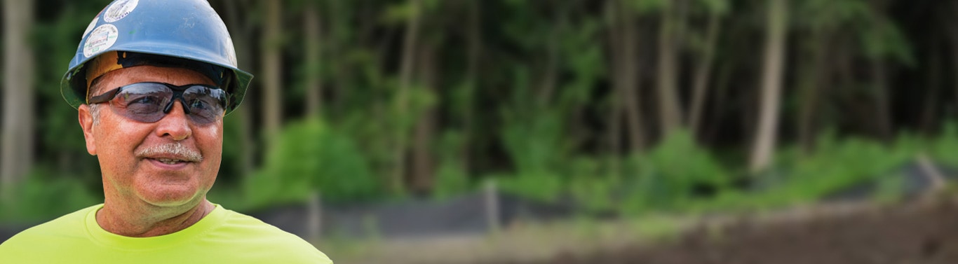 Person wearing a hard hat and high-visibility shirt stands outdoors near a wooded construction area.