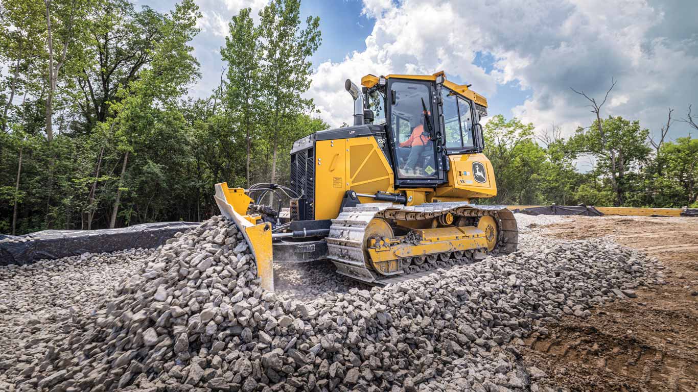 Tracked bulldozer pushes a pile of crushed rock at an active construction site surrounded by trees.