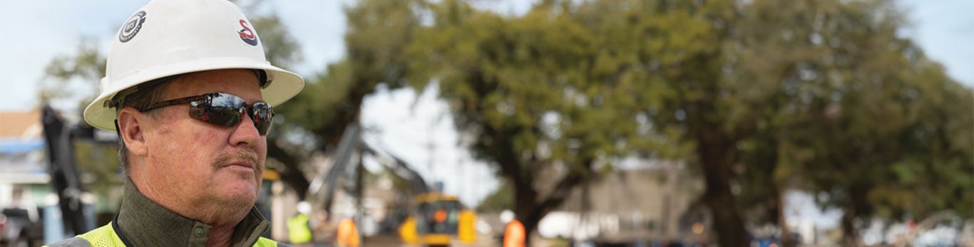 Evan wearing a hard hat and sunglasses standing in front of the job site