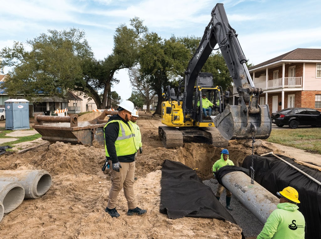 Two workers manually guide a large pipe being lowered into a trench by an excavator