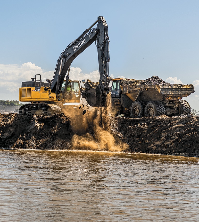 John Deere 470G Excavator digging into the soil next to a body of water and loading it into a John Deere Articulated Dump Truck.