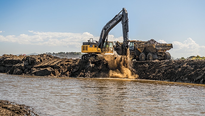 A John Deere 470G LC Excavator is moving a bucket full of dirt from a lagoon to be dumped into a 410E II Articulated Dump Truck.