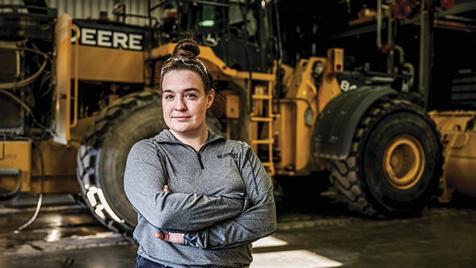 Michelle Roth stands in front of a large wheel loader being worked on.