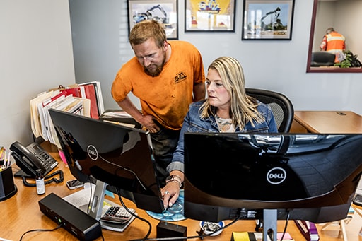 Administrative Assistant Melissa Cordner works at her desk while Mike Bertrand looks over her shoulder. 