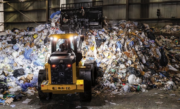 A John Deere 644X Tier Wheel Loader lifts its bucket, moving recyclable items up a pile.