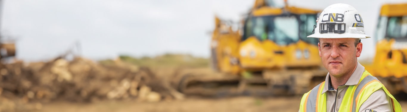 Worker in safety gear at a dirt-covered site with yellow bulldozers in the background