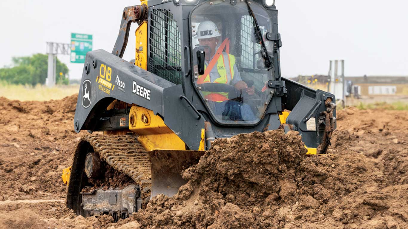 Worker in safety gear operating a John Deere loader on a muddy construction site.