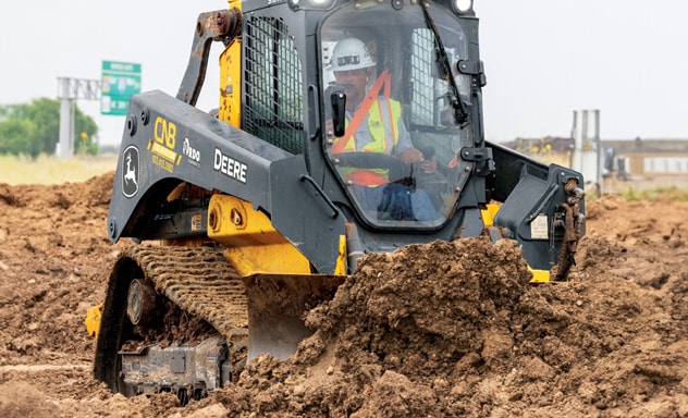 Worker in safety gear operating a John Deere loader on a muddy construction site.