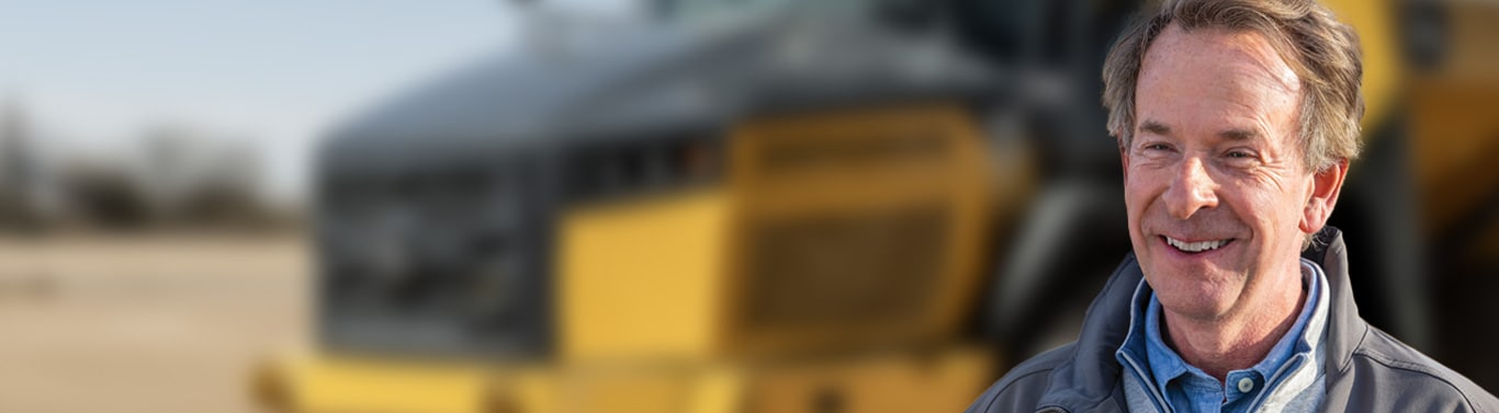 Smiling man stands in front of a large yellow construction vehicle.