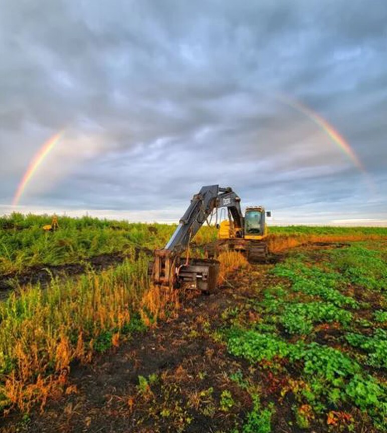 John Deere Compact Excavator digging with a rainbow arching over the machine.