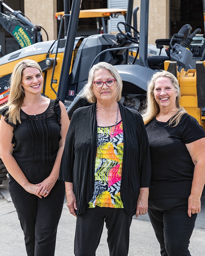 Representing three generations, Amanda Matson, Janet Ferguson, and Nikki Worden of Newman Backhoe Service stand in front of a John Deere 310L Backhoe.