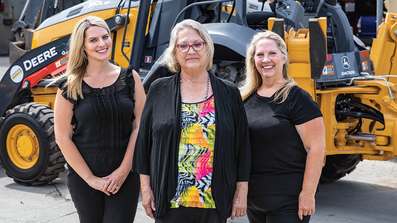 Amanda Matson, Janet Ferguson, and Nikki Worden of Newman Backhoe Service stand in front of a John Deere 310L Backhoe.