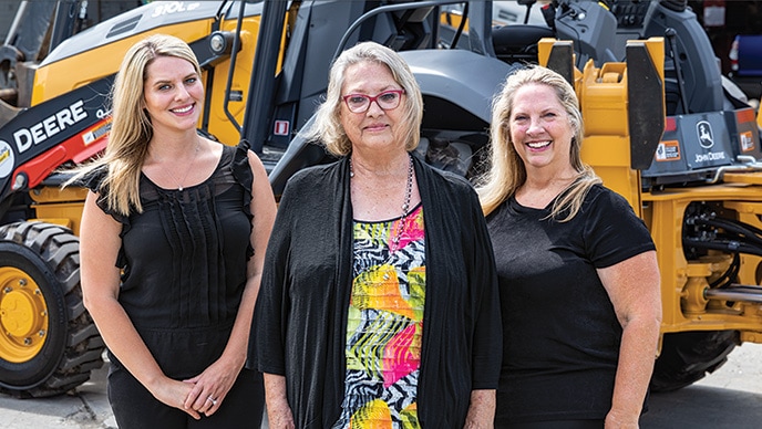 Amanda Matson, Janet L Ferguson, and Nikki J Worden of Newman Backhoe Service stand in front of a John Deere 310L Backhoe.