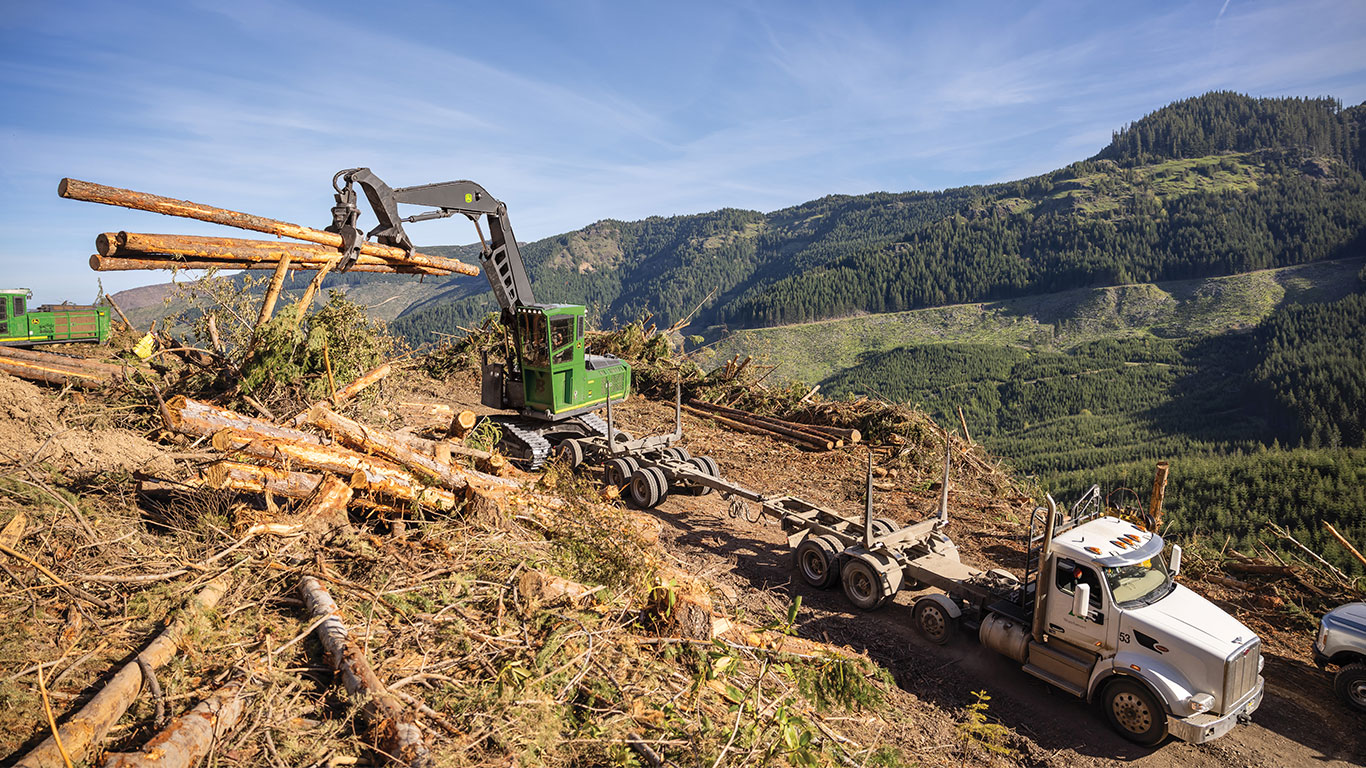 A logging machine unloads tree logs onto a truck on a cleared hillside, surrounded by dense forested mountains under a clear blue sky.