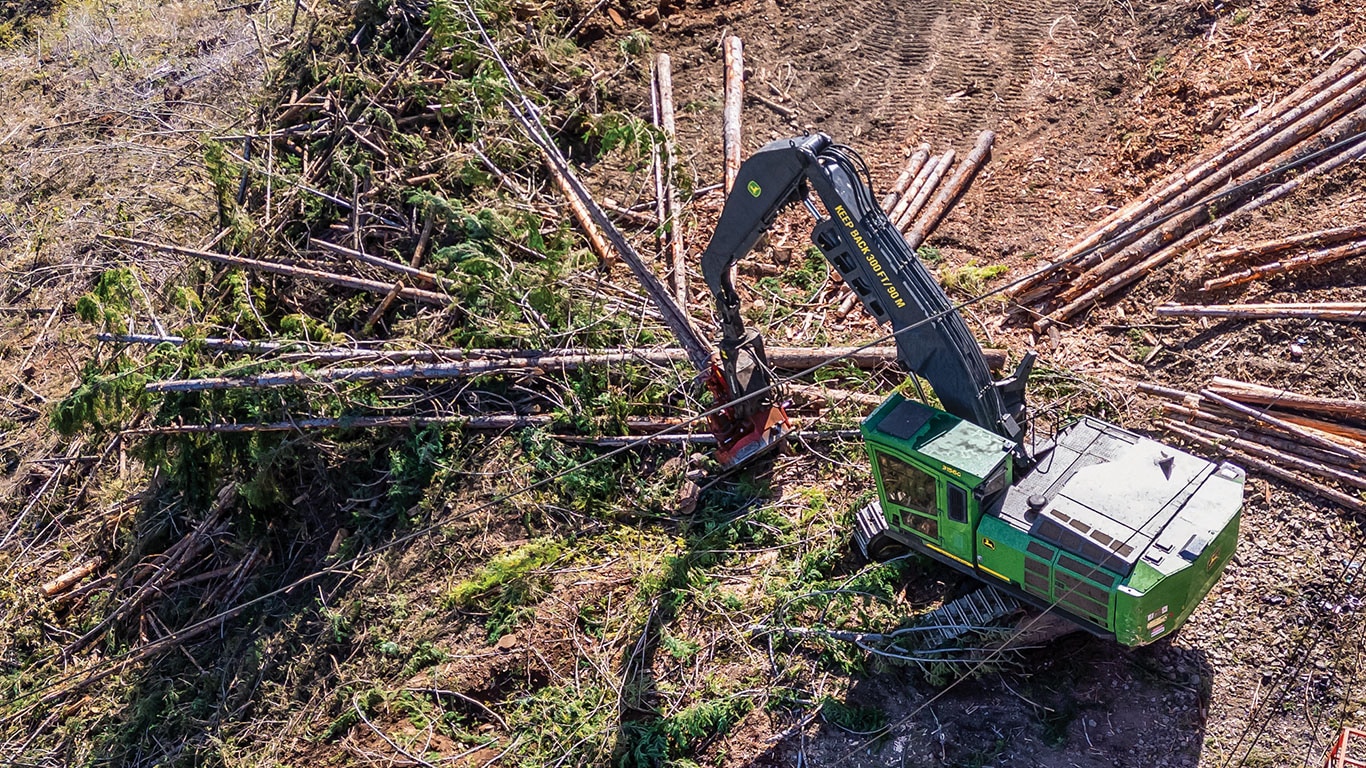 Aerial view of a logging machine with an extended arm, processing timber on a forested site surrounded by cut trees.