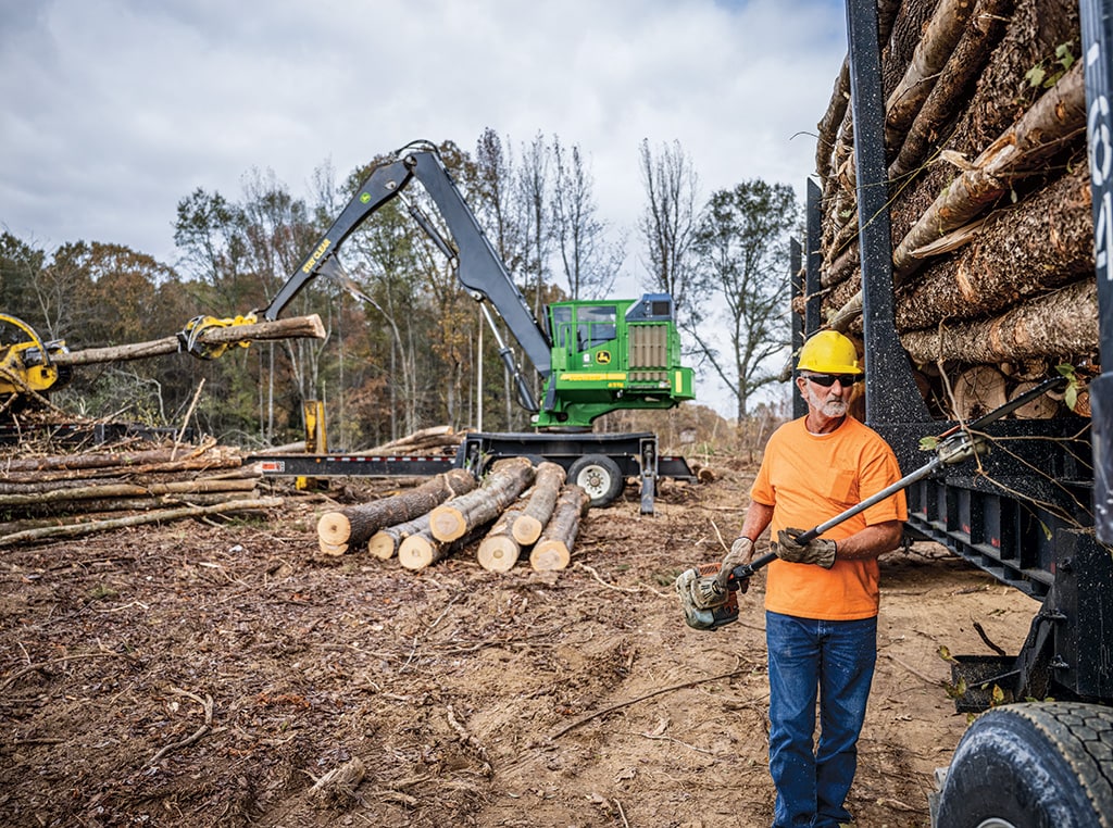 Terry trims limbs from logs on a trailer while the 437E Knuckleboom Loader pulls trees from the delimber.