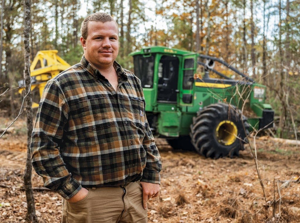 Ryan stands in the woods with his company’s 843L-II Wheeled Feller Buncher with its cab door open behind him.