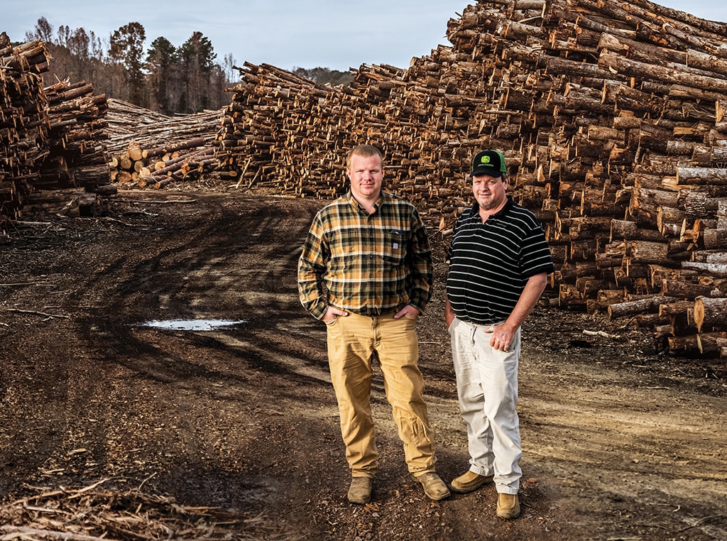 Ryan poses to the left of Edwin on the logging road with large piles of stacked hardwood on both sides.