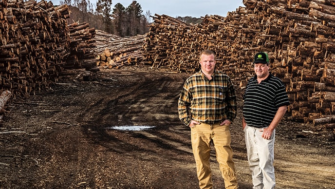 Son Ryan poses to the left of his dad Edwin on the logging road with stacked hardwood on both sides of them at the Edwin Taylor & Son Logging woodyard in Duck Hill, Mississippi.